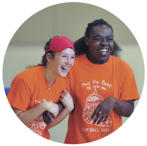 A candid photo of two teens laughing during Beep Ball. They are both wearing orange tshirts with the event logo on it.