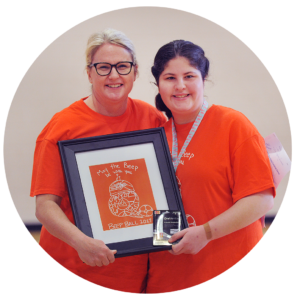 Posed photo of Nancy the Executive Director awarding the 2017 Beep Ball Prize to a young woman. They are both holding a framed logo of Beep Ball in front of them.