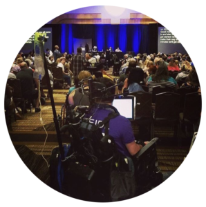 Candid photo taken during a meeting at the back of the room - a variety of men and women are sitting in seats facing the main podium where the speakers are sitting with closed captioning screens to the right and left. The closest person to the camera is sitting in a wheelchair.