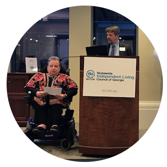 Candid photo of presenters at a Georgia SILC meeting. A woman sitting in her wheelchair is to the left looking at the paper she is holding and a man is standing behind a podium with a white sign on the podium that reads "Statewide Independent Living Council of Georgia"