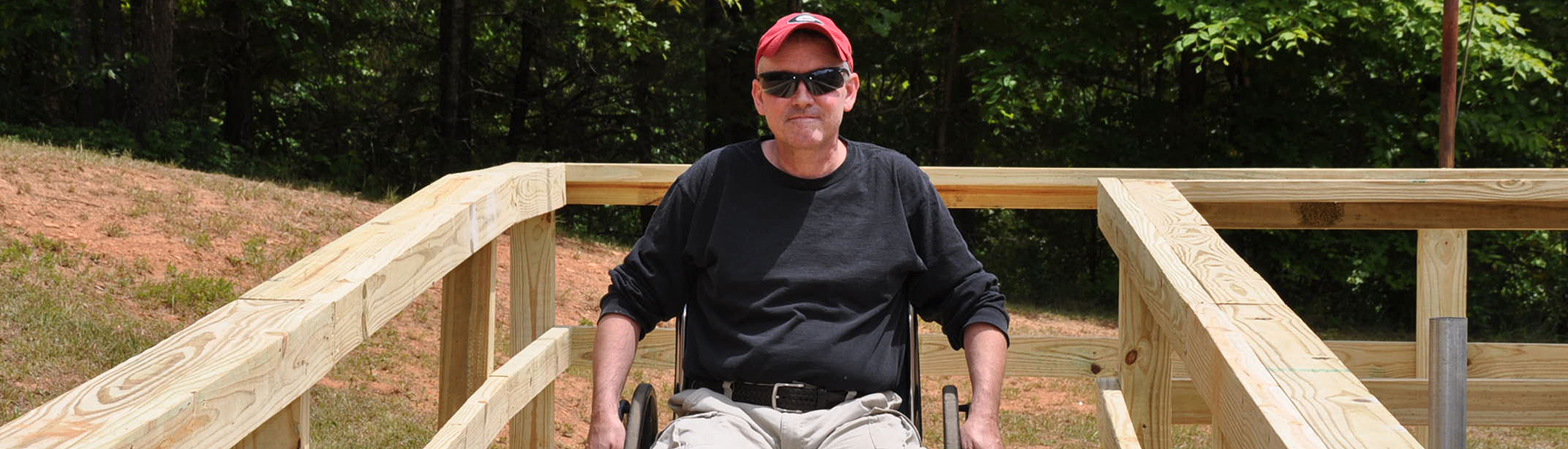 A man in a wheelchair, sunglasses and baseball cap rolls down a newly built home ramp.