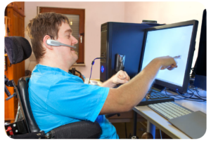 A man sitting in his wheelchair is pointing at a computer screen on his desk. He appears to be speaking into the headset positioned on his ear.