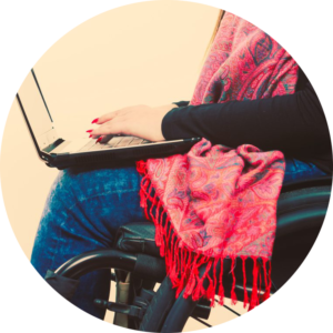 Circle photo: a person using a laptop while sitting in her wheelchair. The focu of the photo is the laptop and the hands using the laptop.
