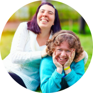 Circle photo: two young ladies are outside laughing. The woman on the right is laying on the ground and propping her head in her hands while the woman on the right leans against the other with her hands on her shoulders.